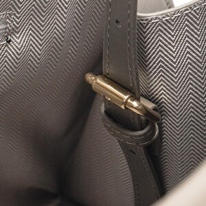 May include: Close-up of a gray handbag interior with a herringbone pattern lining. A gray leather strap with a gold-colored metal clasp is visible. The bag's interior is a lighter shade of gray.