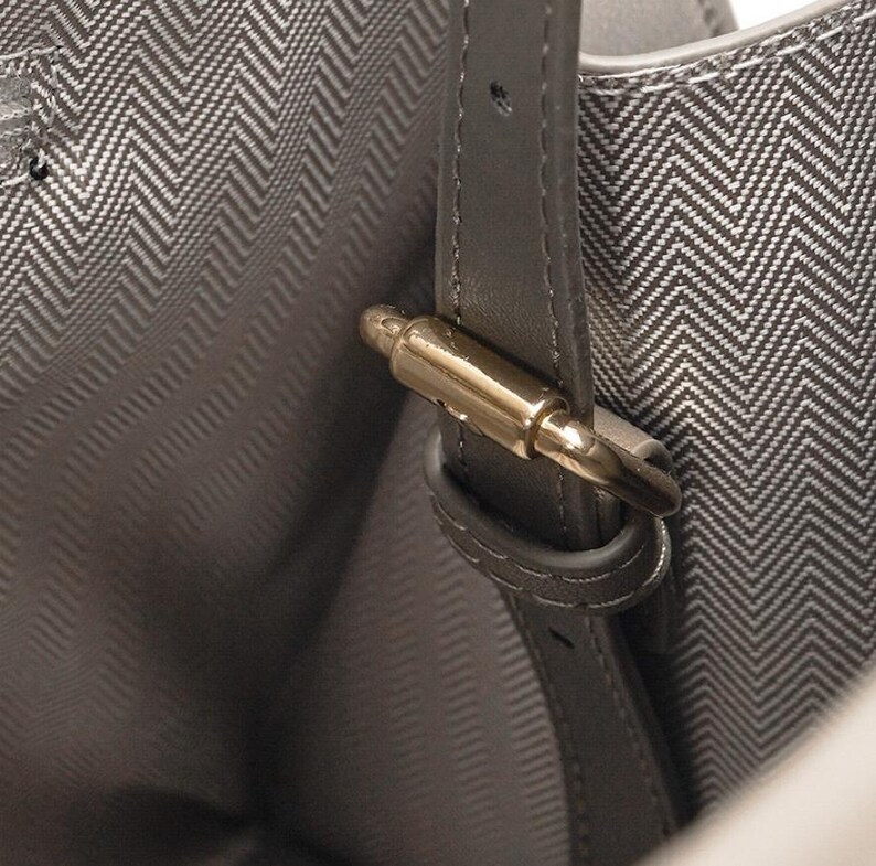 May include: Close-up of a gray handbag interior with a herringbone pattern lining. A gray leather strap with a gold-colored metal clasp is visible. The bag's interior is a lighter shade of gray.