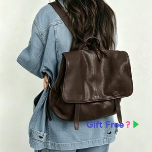 Könnte beinhalten: Ein dunkelbrauner Lederrucksack mit einer großen Umschlagklappe, zwei vertikalen Riemen und einem Tragegriff. Der Rucksack wird über einer hellblauen Jeansjacke getragen. Der Text "Gift Free ?" ist in der unteren rechten Ecke zu sehen.