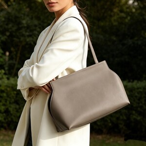 May include: A taupe-colored handbag with a structured design and a single shoulder strap. The bag has a rectangular shape with curved edges and a contrasting white stitch detail. The bag is held by a person wearing a cream-colored blazer.