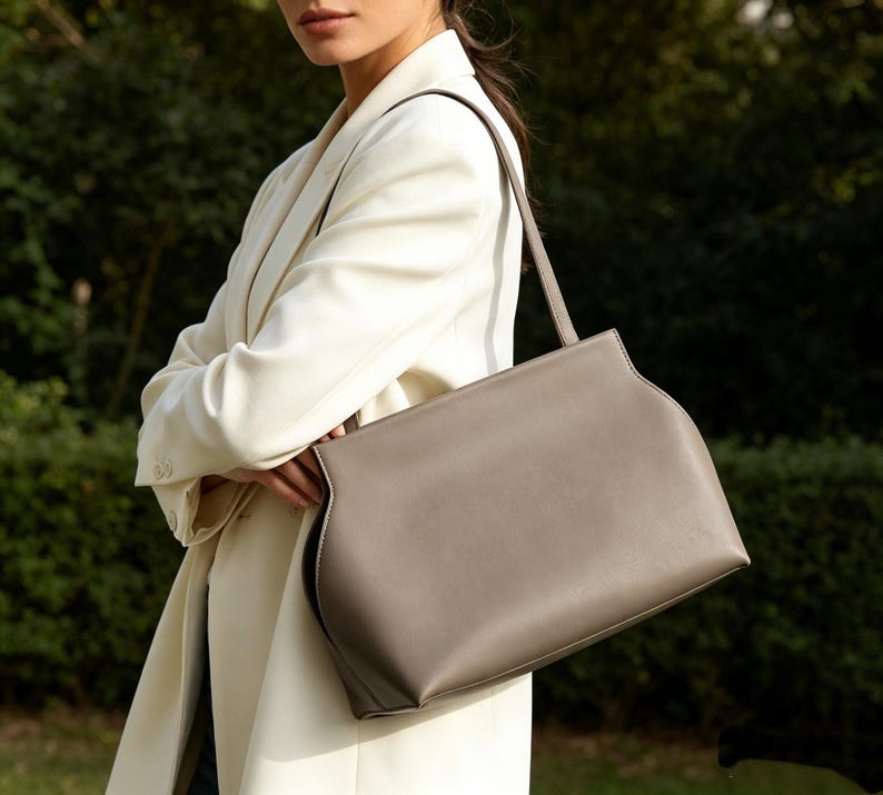 May include: A taupe-colored handbag with a structured design and a single shoulder strap. The bag has a rectangular shape with curved edges and a contrasting white stitch detail. The bag is held by a person wearing a cream-colored blazer.