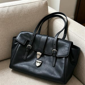 May include: A black leather handbag with a flap closure and silver-tone hardware. The bag has two top handles and adjustable straps with buckles. The bag is on a beige sofa.