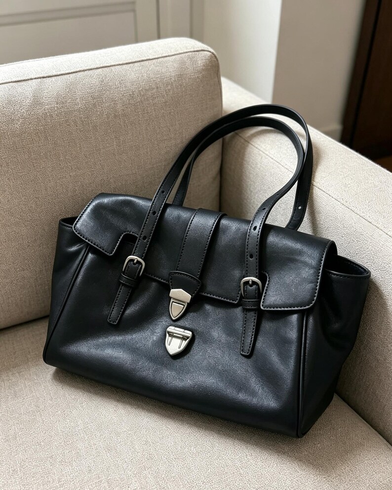 May include: A black leather handbag with a flap closure and silver-tone hardware. The bag has two top handles and adjustable straps with buckles. The bag is on a beige sofa.