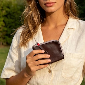 May include: A small, dark brown leather zippered pouch with a smooth finish. The pouch features a light grey fabric gusset along the top edge and a thin leather zipper pull accented with a small, bright red thread detail at the base of the pull.