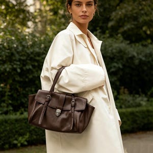 May include: A brown leather handbag with a flap closure and silver-tone hardware. The bag has two buckle straps on the front and a top handle. The woman is wearing a cream-coloured trench coat.
