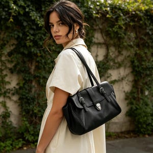 May include: Black leather satchel bag with silver-tone hardware. The bag has a flap closure with two buckle straps and a central clasp. The bag has a structured shape and a long shoulder strap. The background is a green wall of foliage.