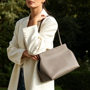 May include: A light gray handbag with a shoulder strap. The bag has a rectangular shape with curved edges and a subtle seam detail. The bag is held by a person wearing a cream-colored blazer.