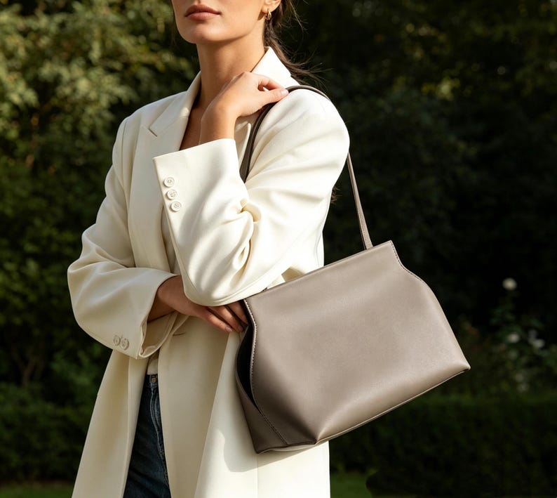 May include: A light gray handbag with a shoulder strap. The bag has a rectangular shape with curved edges and a subtle seam detail. The bag is held by a person wearing a cream-colored blazer.