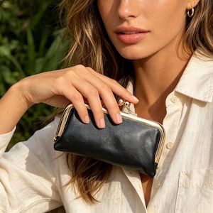 May include: A rectangular black leather clutch purse featuring a gold-toned metal frame with a kiss-lock clasp closure. The purse has a smooth, slightly textured surface and a structured shape, designed with a metallic gold hinge mechanism at the top.
