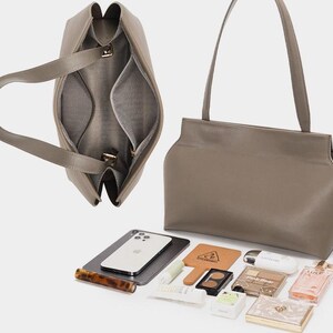 May include: A taupe-colored leather handbag with a structured design, featuring a top handle and a spacious interior. The bag is open, revealing internal compartments. Various beauty products, a smartphone, and a small mirror are displayed beside the bag.