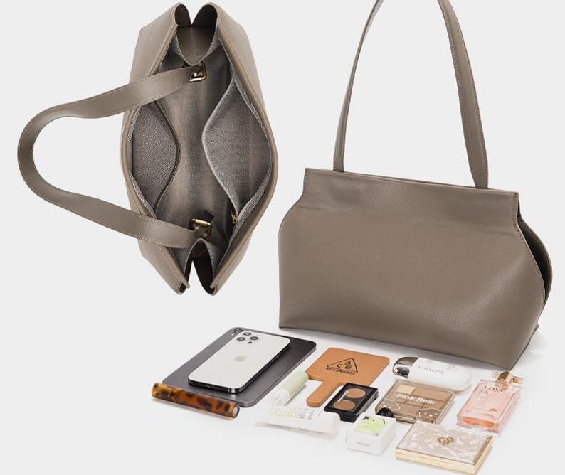 May include: A taupe-colored leather handbag with a structured design, featuring a top handle and a spacious interior. The bag is open, revealing internal compartments. Various beauty products, a smartphone, and a small mirror are displayed beside the bag.
