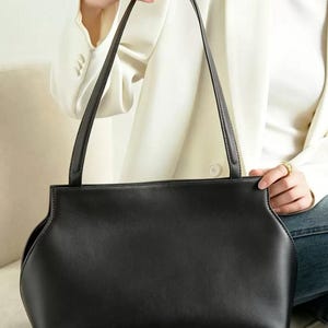 May include: A black handbag with long, thin straps. The bag has a rectangular shape with curved sides and a smooth texture. The straps are also black and appear to be made of the same material as the bag. The background shows a person wearing a white blazer.