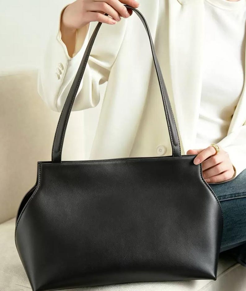 May include: A black handbag with long, thin straps. The bag has a rectangular shape with curved sides and a smooth texture. The straps are also black and appear to be made of the same material as the bag. The background shows a person wearing a white blazer.