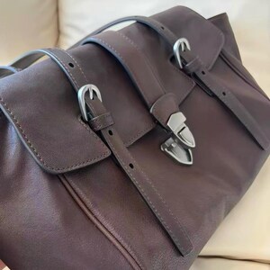 May include: A brown leather satchel bag with silver buckles and straps. The bag features a flap closure and a structured design. The leather has a matte finish. The bag is designed for carrying items.