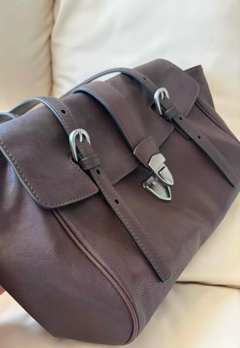 May include: A brown leather satchel bag with silver buckles and straps. The bag features a flap closure and a structured design. The leather has a matte finish. The bag is designed for carrying items.