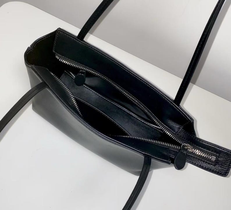 以下が含まれることがあります： 細いショルダーストラップと銀色の金属製ジッパーが付いた黒いトートバッグ。内部には中央にジッパー付きの仕切りがあり、メインスペースが2つのセクションに分かれています。バッグ全体に滑らかな黒い裏地が付いています。