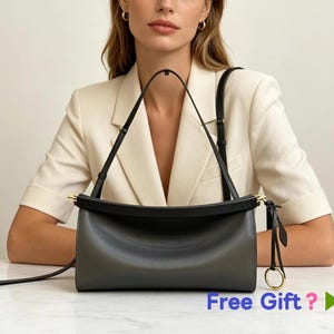 Puede incluir: Un bolso de hombro horizontal y rectangular de color gris oscuro con acabado liso, borde superior negro y herrajes dorados. El bolso cuenta con una correa de hombro negra fina y ajustable y un detalle de anillo dorado circular en la cremallera.