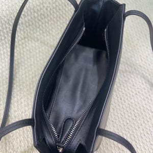 以下が含まれることがあります： 細いストラップと上部にファスナーが付いた黒いショルダーバッグ。内側は黒い素材で裏打ちされており、側面にファスナー付きポケットがあります。バッグは滑らかなエッジを持つ構造的な長方形の形状をしています。