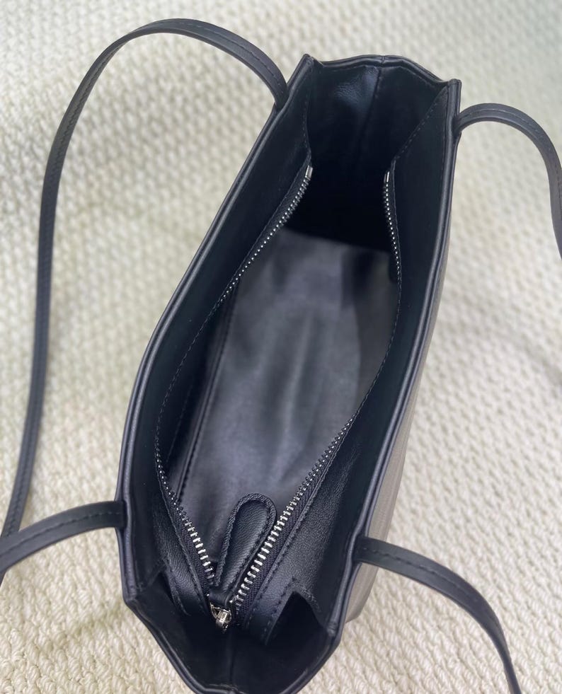 以下が含まれることがあります： 細いストラップと上部にファスナーが付いた黒いショルダーバッグ。内側は黒い素材で裏打ちされており、側面にファスナー付きポケットがあります。バッグは滑らかなエッジを持つ構造的な長方形の形状をしています。
