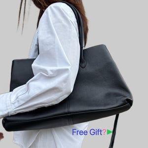 Puede incluir: Un bolso tote grande de cuero negro texturizado con dos correas para el hombro y una costura vertical en la parte delantera. El bolso cuenta con una tira de cuero larga y fina que cuelga del lateral. El texto Free Gift? con flecha verde.