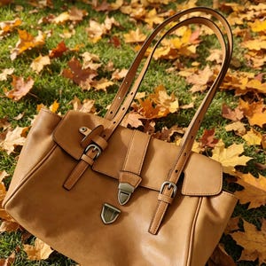 May include: A tan leather handbag with a flap closure and adjustable straps. The bag features silver-coloured buckles and a metal clasp. The bag is on grass and surrounded by autumn leaves.