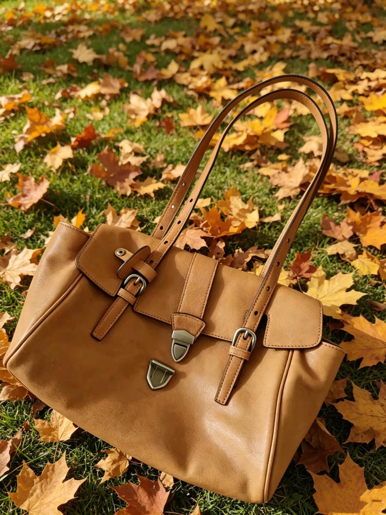 May include: A tan leather handbag with a flap closure and adjustable straps. The bag features silver-coloured buckles and a metal clasp. The bag is on grass and surrounded by autumn leaves.