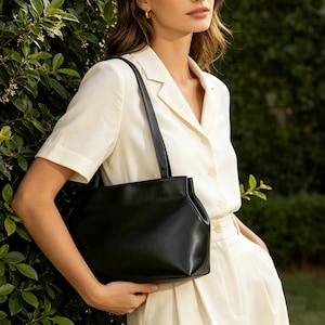 May include: A black leather tote bag with a rectangular shape and a shoulder strap. The bag is held by a person wearing a cream-colored button-down shirt and pants. The background features green foliage.