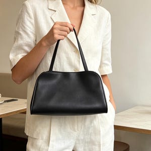 Puede incluir: Un bolso de mano negro con un acabado liso y mate y una forma rectangular estructurada. El bolso cuenta con una sola asa superior delgada y un borde superior curvado, ofreciendo una estética limpia para llevar objetos.