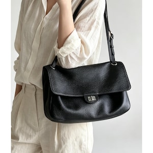 May include: A black leather shoulder bag with a pebbled texture, featuring a rectangular flap, a silver-toned metal turn-lock closure, and an adjustable shoulder strap with silver hardware, worn over the shoulder.