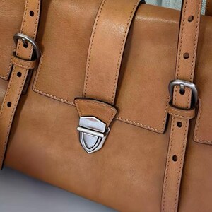 May include: A tan leather satchel bag with silver buckles and straps. The bag has a flap closure with a silver clasp. The leather is smooth and the stitching is visible. The bag is a classic design.