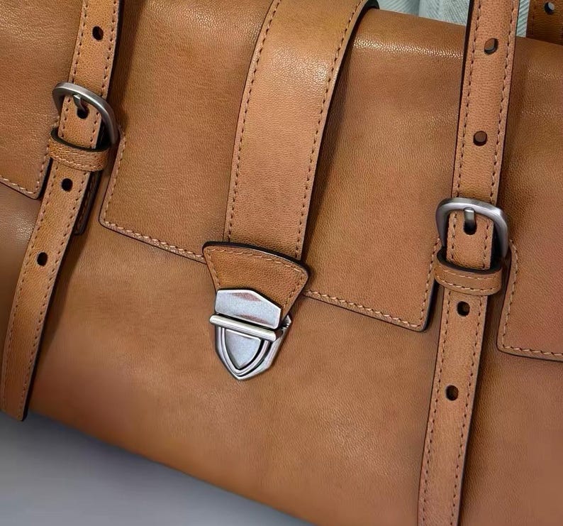 May include: A tan leather satchel bag with silver buckles and straps. The bag has a flap closure with a silver clasp. The leather is smooth and the stitching is visible. The bag is a classic design.