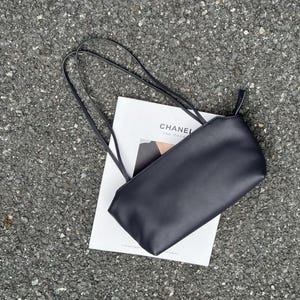 以下が含まれることがあります： 黒い長方形のショルダーバッグで、2本の細いストラップが付いています。白い長方形のカードの上に置かれており、カードには黒い文字で「CHANEL FINE JEWELRY」と書かれています。バッグは滑らかでマットな質感で、金具が見えないシンプルでクリーンなデザインです。