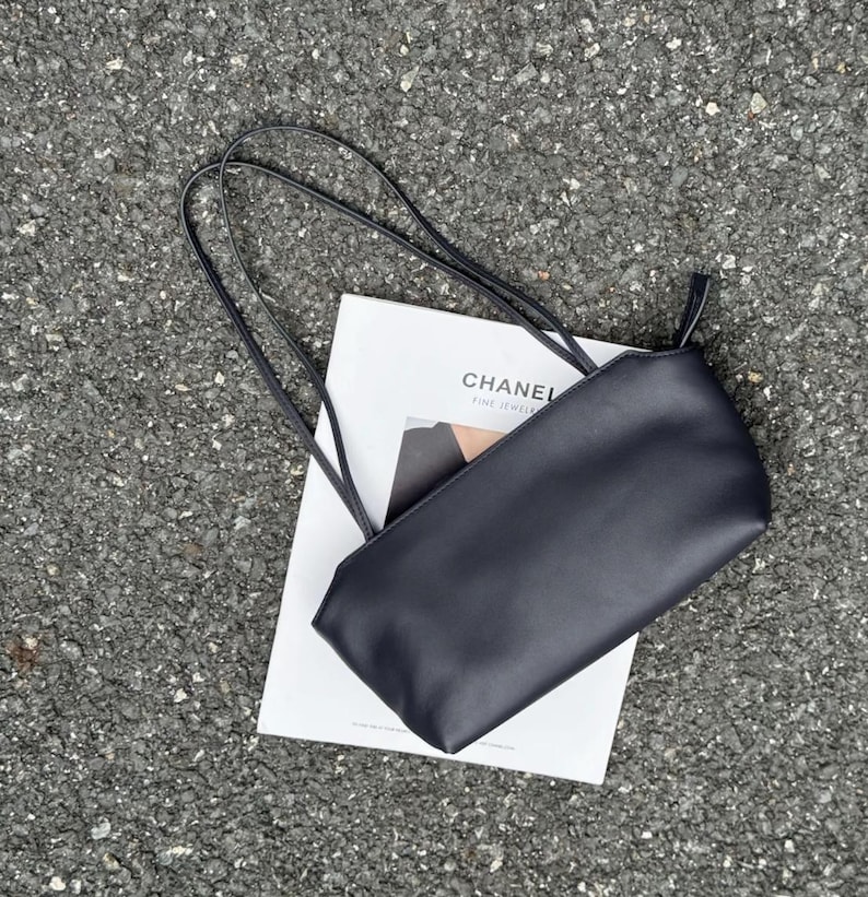 以下が含まれることがあります： 黒い長方形のショルダーバッグで、2本の細いストラップが付いています。白い長方形のカードの上に置かれており、カードには黒い文字で「CHANEL FINE JEWELRY」と書かれています。バッグは滑らかでマットな質感で、金具が見えないシンプルでクリーンなデザインです。