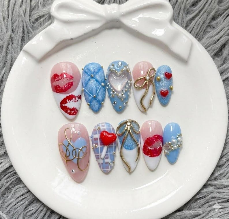 May include: A set of ten artificial nails with various designs, including red lips, blue quilted patterns, heart accents, and gold bow details. The nails are arranged on a white, bow-shaped plate.