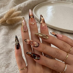May include: Long almond-shaped artificial nails featuring a mix of designs including glossy brown tortoiseshell patterns, metallic gold liquid-like accents, hand-painted beige floral motifs, and small embedded clear rhinestones on a nude base.