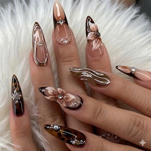 May include: A set of long, almond-shaped acrylic nails with various designs. Some nails feature a tortoiseshell pattern, while others have floral accents and metallic embellishments. The color palette includes shades of brown, nude, and clear.