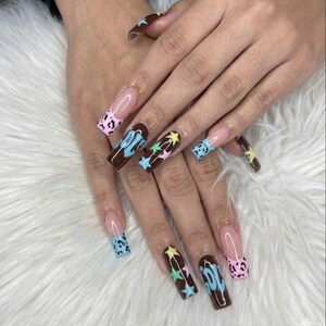 May include: A set of long, square-shaped acrylic nails. Some nails are painted a dark brown with star and swirl designs in pastel colors. Others have a pink leopard print or a French tip with a clear base.