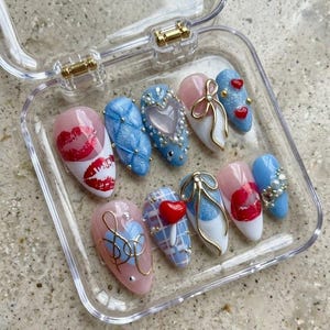 May include: A set of ten artificial nails in a clear plastic case. The nails feature various designs, including red lips, blue quilted patterns, hearts, bows, and gold accents. The nails are in shades of pink, white, and blue.