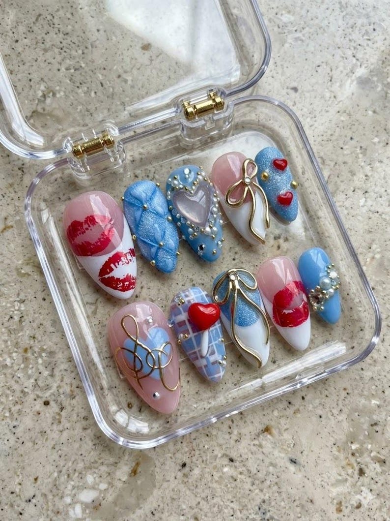 May include: A set of ten artificial nails in a clear plastic case. The nails feature various designs, including red lips, blue quilted patterns, hearts, bows, and gold accents. The nails are in shades of pink, white, and blue.
