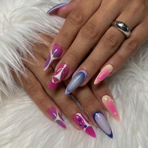 May include: A close-up of a hand with long, pointed, and colorful acrylic nails. The nails feature a variety of designs, including gradients, geometric shapes, and gold accents. The nails are painted in shades of pink, purple, blue, and white. A silver ring is on the finger.