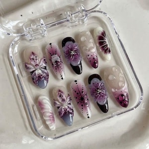 May include: A set of ten artificial nails in a clear plastic case. The nails feature various designs, including 3D purple flower embellishments, dot patterns, and gradient color schemes of white, pink, and purple. Some nails have a black base.