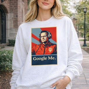 May include: White sweatshirt with a graphic of a man wearing headphones and the text "Google Me." The sweatshirt is paired with a cream-colored baseball cap with a red logo. The person is wearing blue jeans.