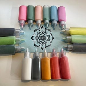 May include: A collection of colorful paint bottles in various shades of pink, green, peach, teal, blue, and yellow. The bottles are arranged around a mandala design on a light blue surface. Additional bottles in white, black, orange, red, and maroon are also present.