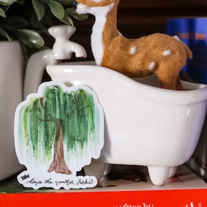 May include: A white ceramic bathtub with a brown and white deer figurine inside. A sticker with a watercolor painting of a weeping willow tree and the text "Here's the weather, Parks" is attached to the side of the tub. The tub is sitting on top of a red book with the text "written by Jessa Hastings" on the cover.
