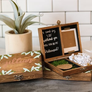 May include: Two wooden boxes with lids. The top box has a chalkboard panel with the text "I can't say 'I do' without you. Will you be my bridesmaid?" The bottom box has a floral design and the name "Jillian" written on it.