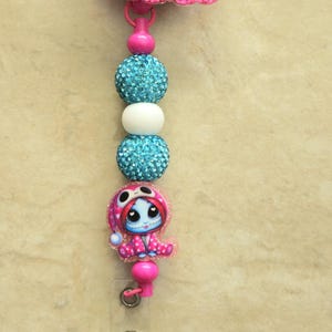 May include: A pink and blue badge reel featuring a cartoon character with red hair and stitched facial features. The reel has a clear plastic strap and a metal clip. Decorative blue and white beads are strung between the character and the reel.