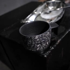 Può includere: Una tazza in ceramica nera con superficie strutturata e un'emblema a croce. La tazza ha un manico quadrato. Un vassoio d'argento è visibile sullo sfondo. Adatta per caffè o tè.