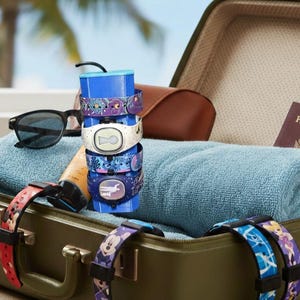 May include: A stack of colorful wristbands with various designs, including floral and character patterns, sits atop a blue base. Sunglasses and a suitcase are in the background, suggesting travel and vacation themes.