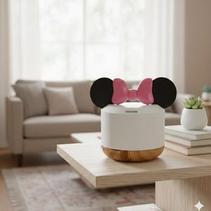 May include: White and wood-tone essential oil diffuser with black ears and a pink bow on top. The diffuser sits on a wooden coffee table in a living room setting. A small succulent plant in a white pot is also on the table.