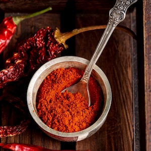 Puede incluir: Un cuenco plateado lleno de vibrante pimiento rojo en polvo, con una cuchara plateada decorativa en su interior. Pimientos rojos secos esparcidos alrededor del cuenco, todo en una bandeja de madera rústica. Primer plano de las especias.